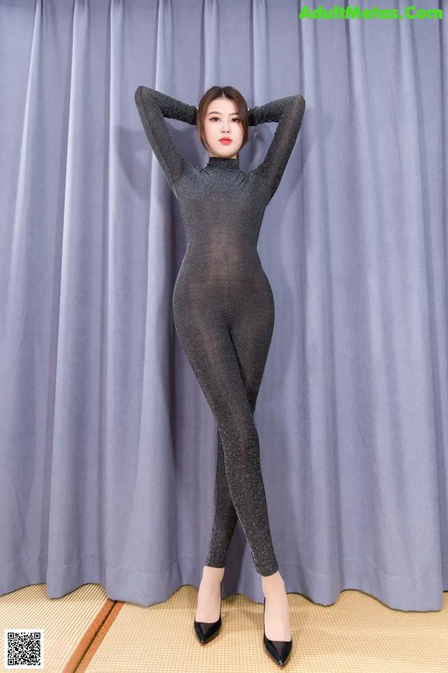A woman in a black bodysuit posing in front of a curtain.